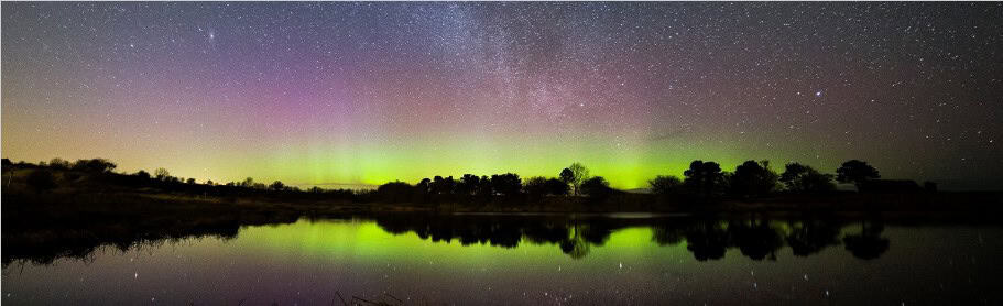 Northern lights glow green and purple above a lake, reflecting the colours and a star-filled night sky.