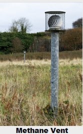 A metal methane vent stands in a grassy field, with another vent visible in the background.